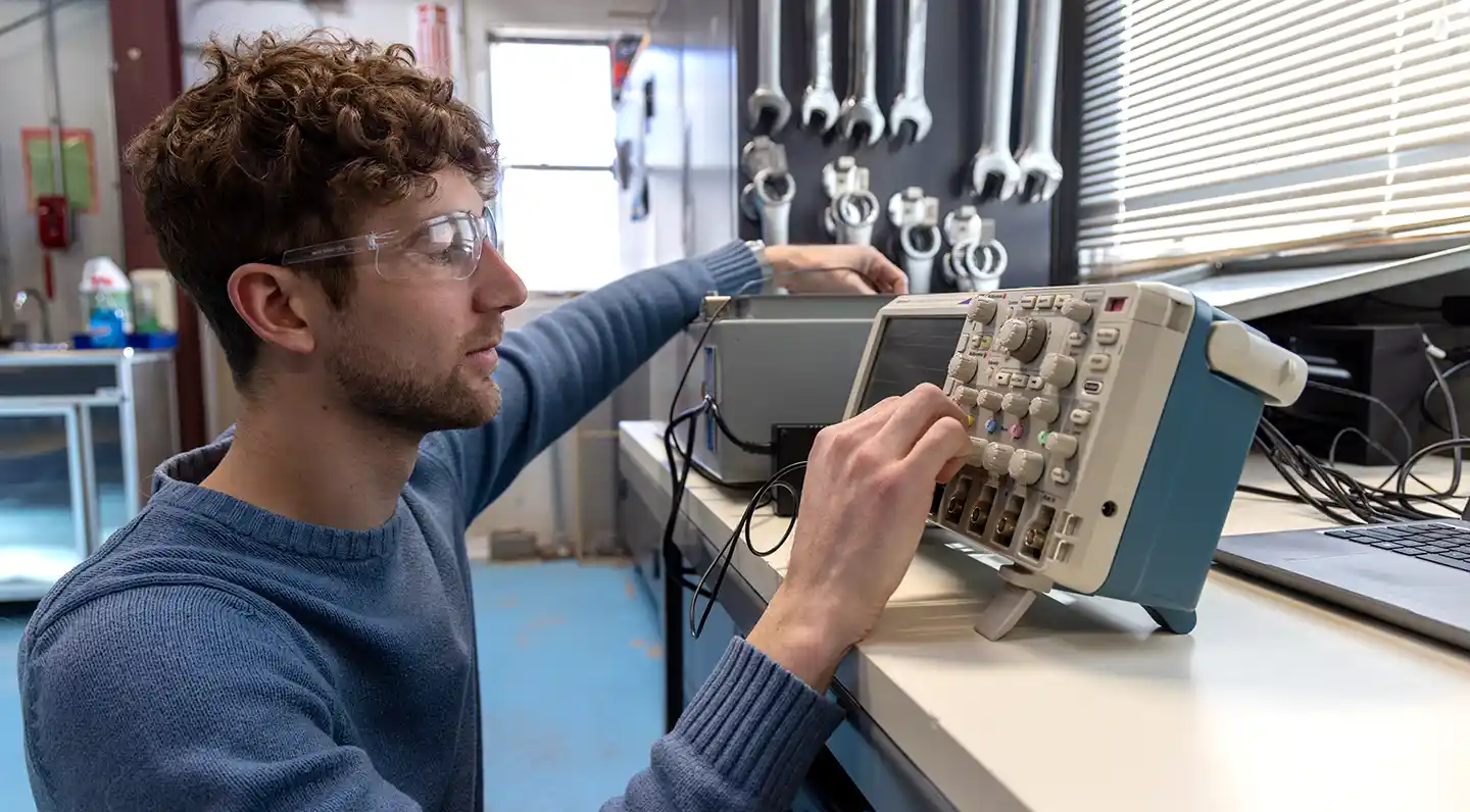 A person wearing safety glasses sitting at a workbench in a workshop, focusing intently as he adjusts the control knobs on a digital oscilloscope.