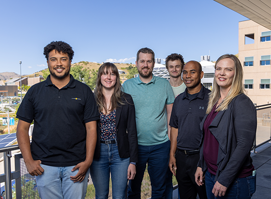Six people smile and pose for a photo while standing on a building patio.