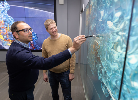 Two people face a glass wall while one draws on it with a marker.