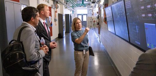 Two people look at a wall with monitors as another stands in front of them and gestures toward the screens.