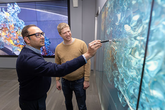 A person using a pen to point at a swirly simulation of combustion gases on a screen, while another person looks on.