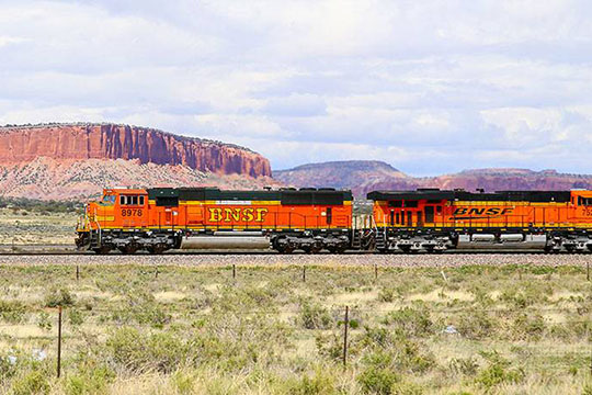 An orange BNSF freight train crosses a flat green plain in front of a colorful desert mesa.
