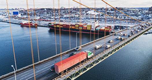 A large semi-truck and other vehicles driving on a suspension bridge over a large body of water.