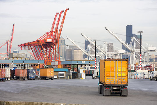 Trucks entering a port, with cranes and a city skyline in the background.