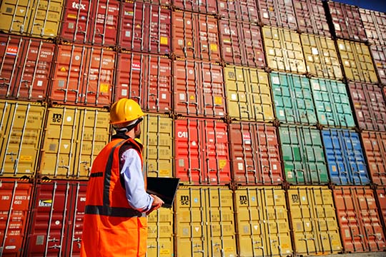A person in a hard hat and safety vest standing in front of colorful shipping containers.