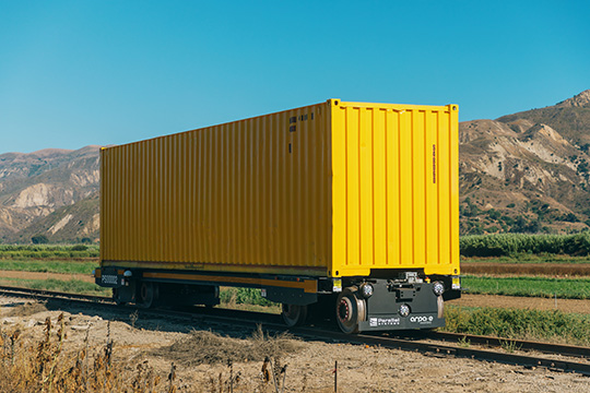 A yellow Parallel Systems autonomous battery-electric rail car against a mountain backdrop