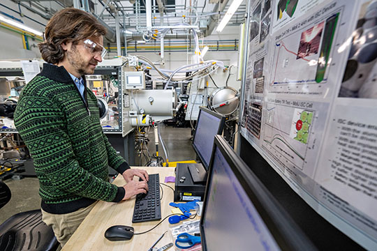 A person standing at a computer with screens in the foreground and battery lab equipment in the background.