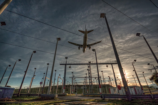 An airplane flying low, photographed from below, with equipment on poles on land in the foreground.