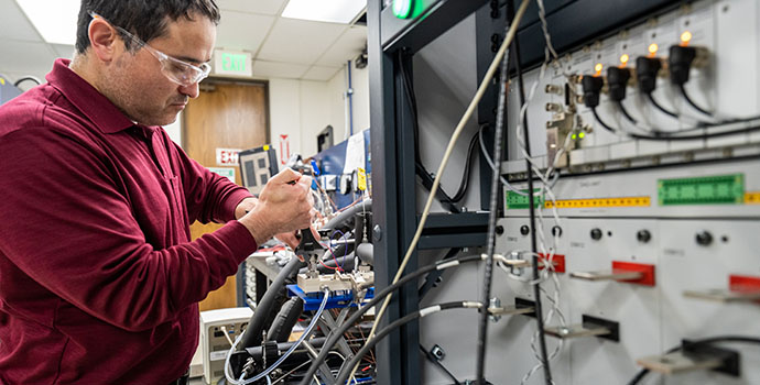 A technician in safety glasses works on complex wired electronic equipment in a laboratory.