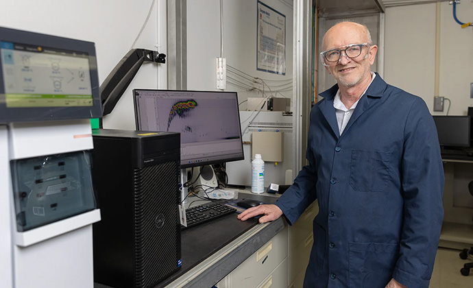 A man wearing a dark blue lab coat stands at a desk with a computer and machine with another screen.